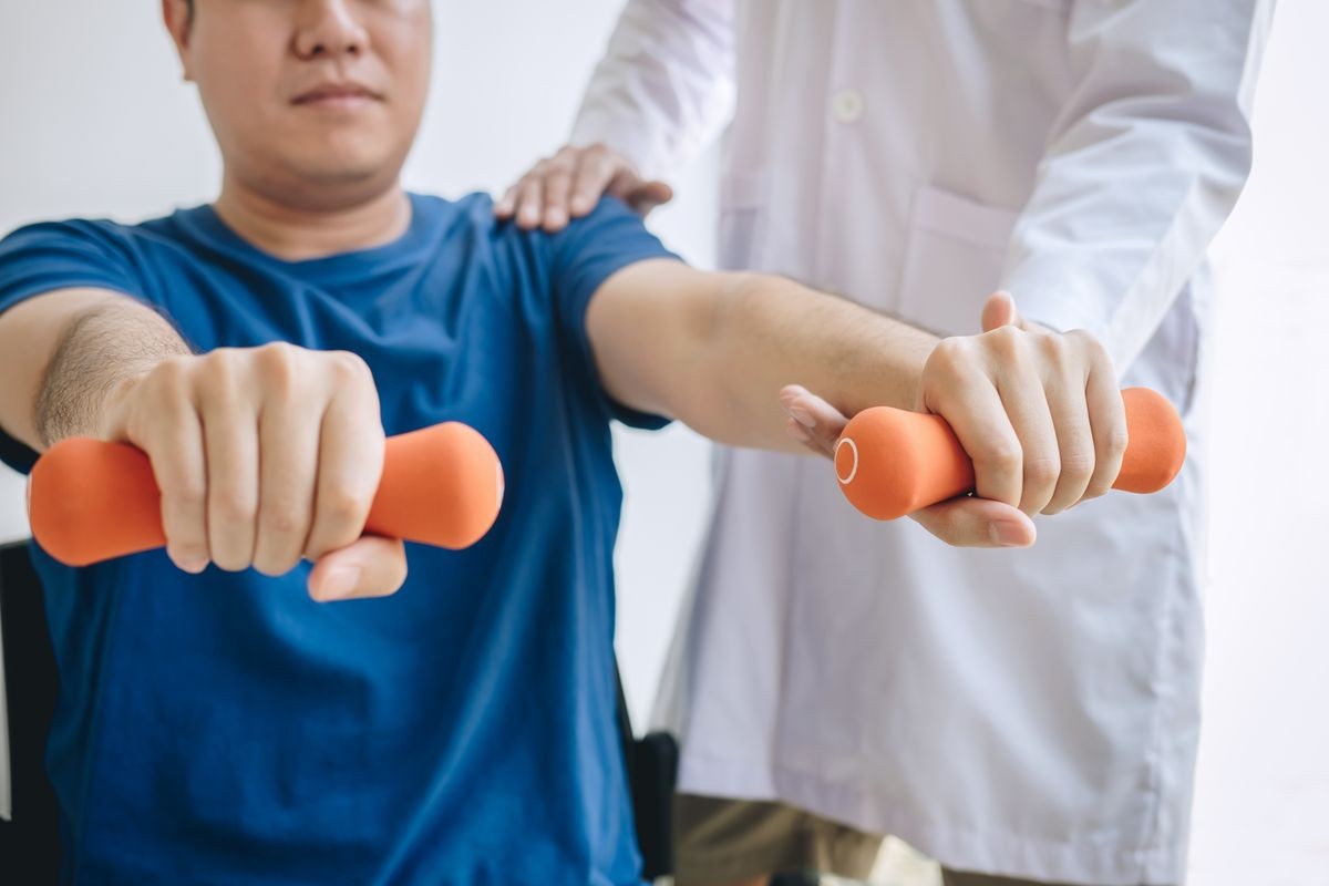 Doctor physiotherapist assisting a male patient while giving exercising treatment on stretching his arm with dumbbell in the clinic, Rehabilitation physiotherapy concept.